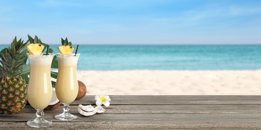 Image of Tasty Pina Colada cocktail on wooden table near ocean, space for text. Banner design