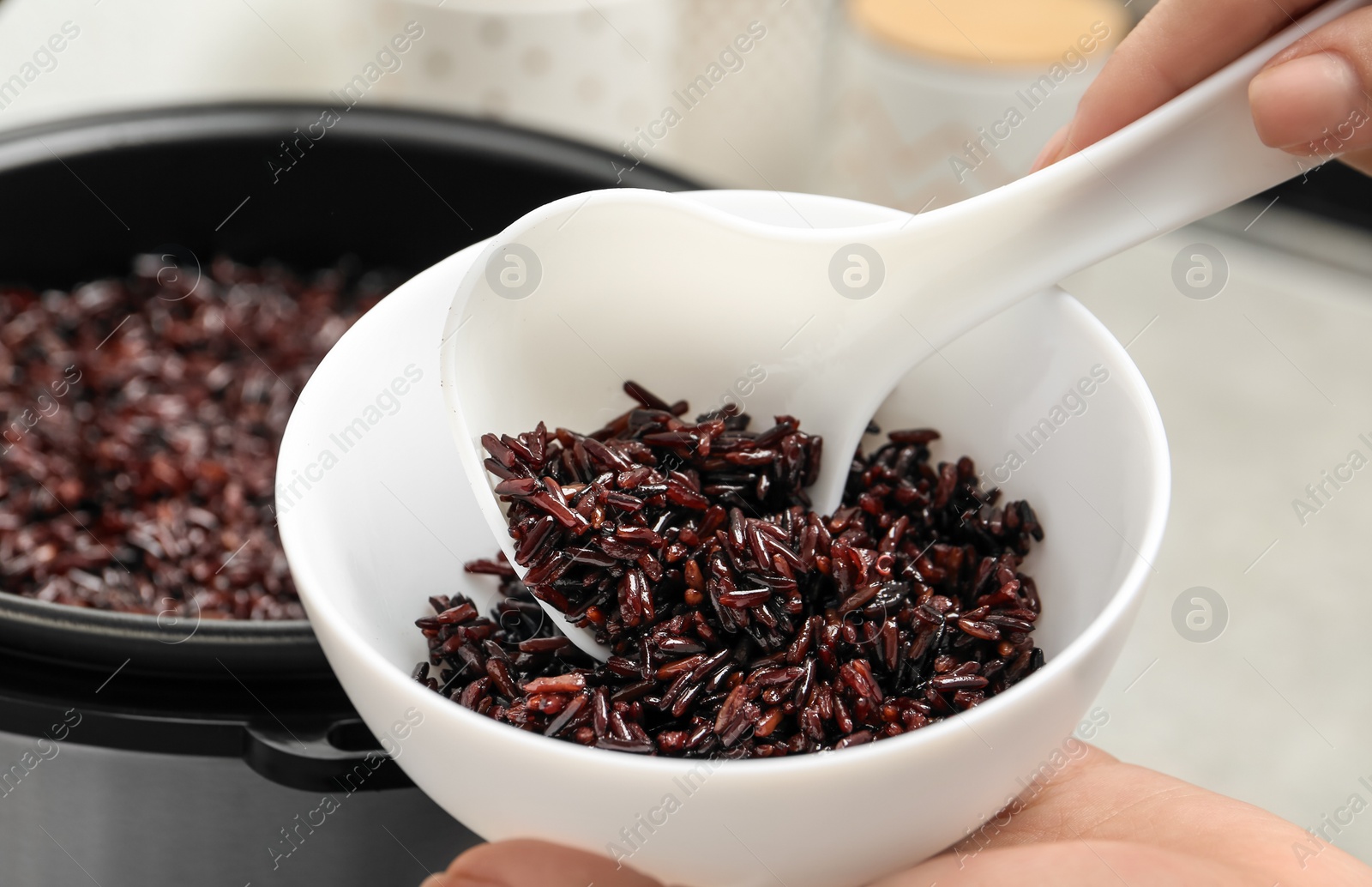 Photo of Woman putting brown rice into bowl from multi cooker in kitchen, closeup