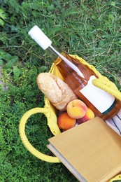 Photo of Yellow wicker bag with book, peaches, baguette and wine on green grass outdoors, top view