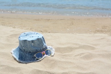 Photo of Jeans hat and sunglasses on sand near sea, space for text. Beach accessories