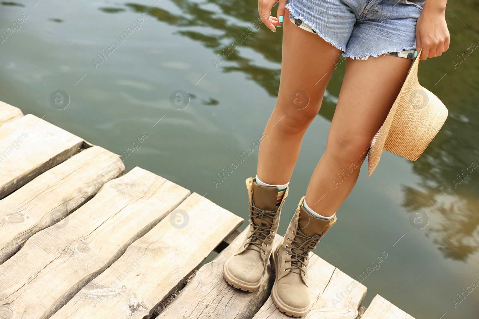 Photo of Young woman with hat on wooden pier near lake, focus on legs. Camping season