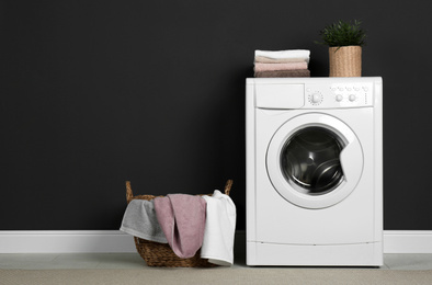 Photo of Modern washing machine with stack of towels, houseplant and laundry basket near black wall. Space for text