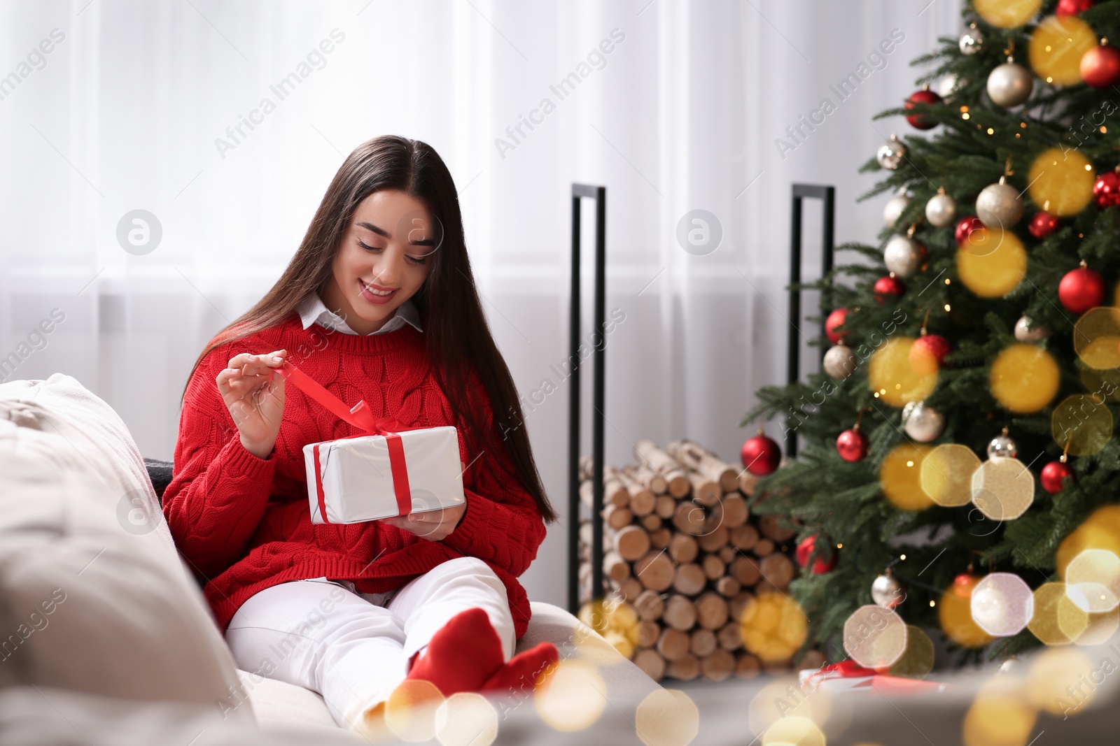 Photo of Smiling woman opening gift near Christmas tree indoors