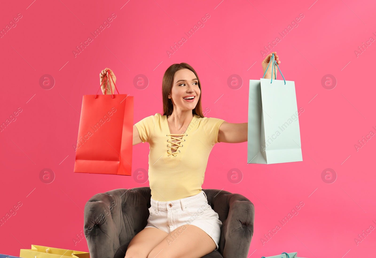 Photo of Beautiful young woman with paper shopping bags in armchair on pink background