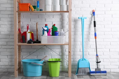 Shelving unit with detergents, cleaning tools and toilet paper near white brick wall indoors