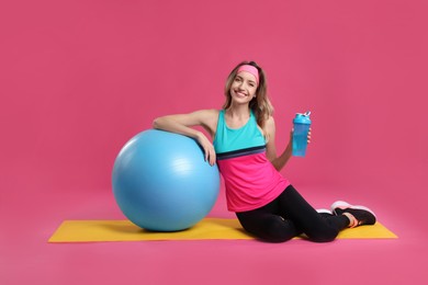 Beautiful woman sitting on yoga mat with fitness ball against pink background