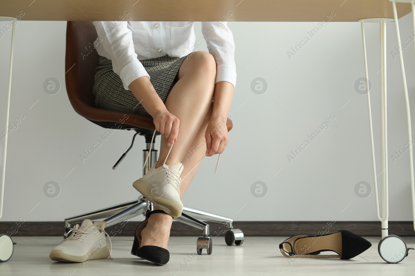 Photo of Woman taking off uncomfortable shoes and putting on sneakers in office, closeup