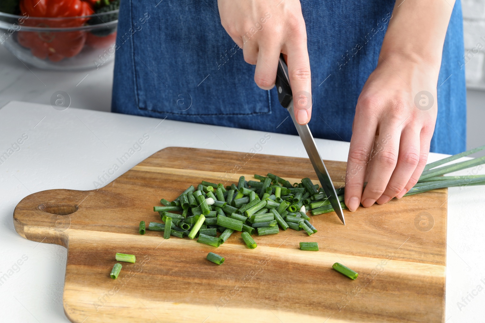 Photo of Woman cutting green spring onion on wooden board at white table, closeup