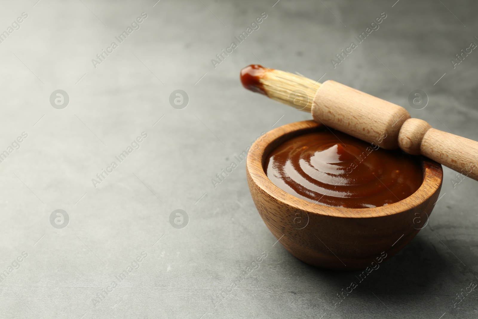 Photo of Tasty barbeque sauce in bowl and brush on grey textured table, closeup. Space for text