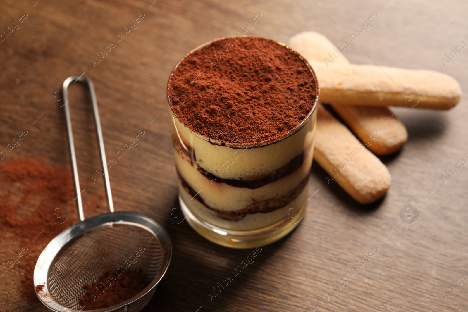 Photo of Delicious tiramisu in glass, biscuits and sieve with cocoa powder on wooden table, closeup