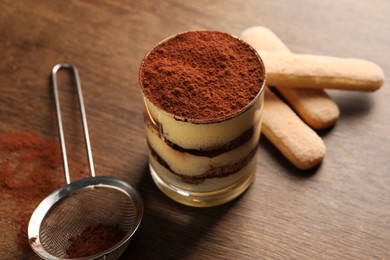 Delicious tiramisu in glass, biscuits and sieve with cocoa powder on wooden table, closeup