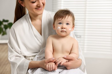 Smiling mother holding her baby with moisturizing cream on face at home, closeup