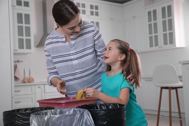 Photo of Young woman and her daughter throwing banana peel into trash bin in kitchen. Separate waste collection