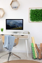 Photo of Green artificial plant wall panel and desk with computer in light room. Interior design