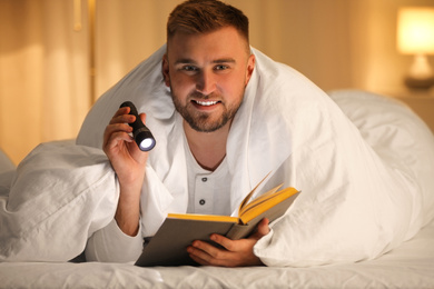 Young man with flashlight reading book in bedroom