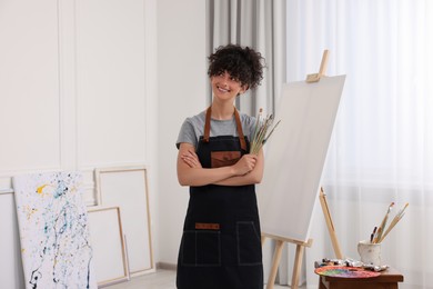 Young woman holding brushes near easel with canvas in studio