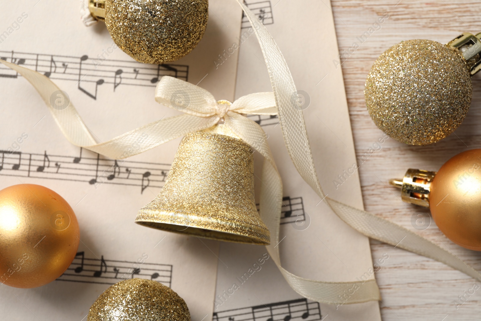 Photo of Golden shiny bell with bow, music sheets and decorative balls on wooden table, flat lay. Christmas decoration