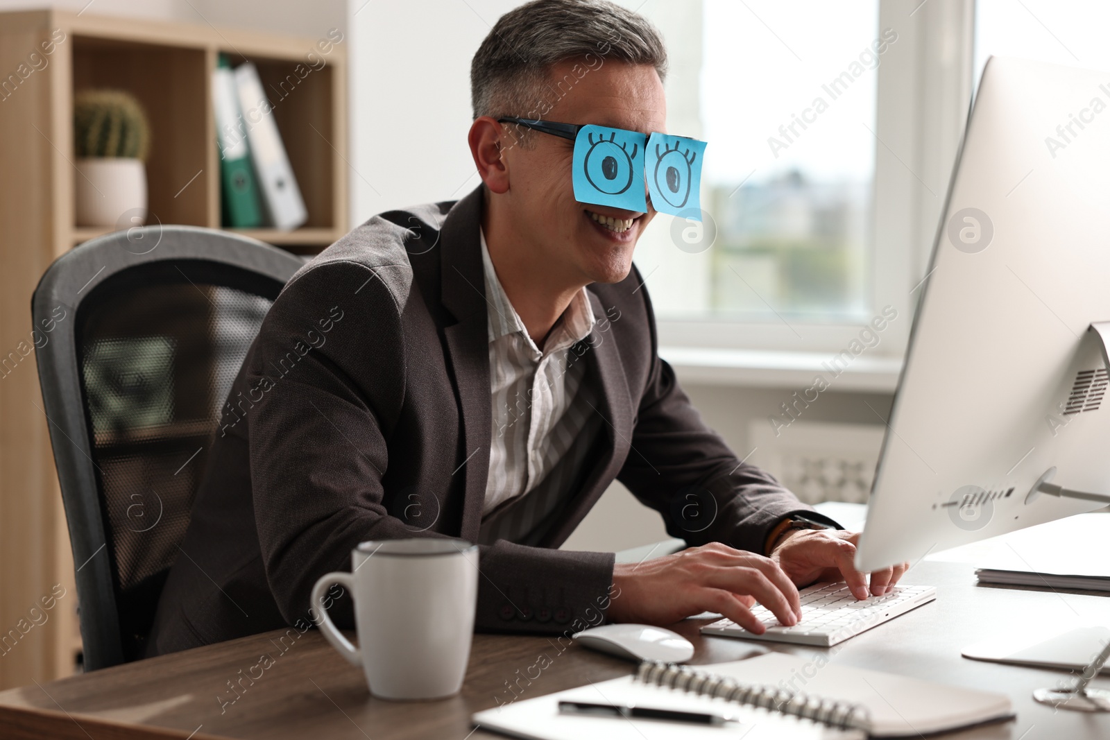 Photo of Man with fake eyes painted on sticky notes working on computer at table in office