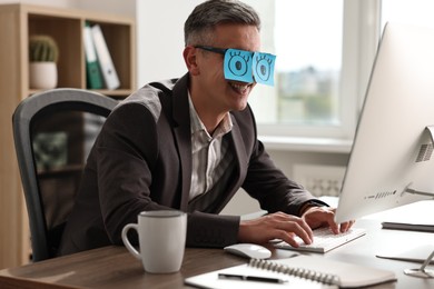 Photo of Man with fake eyes painted on sticky notes working on computer at table in office