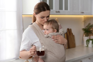 Mother with cup of drink holding her child in sling (baby carrier) in kitchen