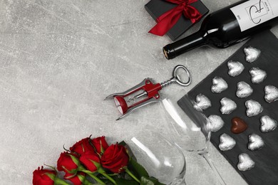 Photo of Bottle of red wine, glasses, heart shaped chocolate candies, corkscrew, roses and gift box on light grey textured table, flat lay. Space for text