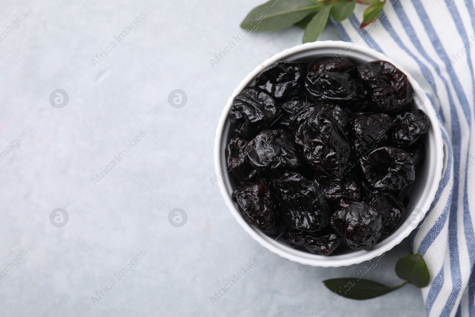 Photo of Sweet dried prunes in bowl and green leaves on light grey table, top view. Space for text