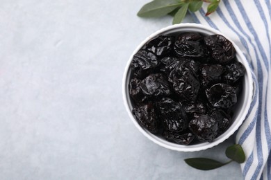 Sweet dried prunes in bowl and green leaves on light grey table, top view. Space for text