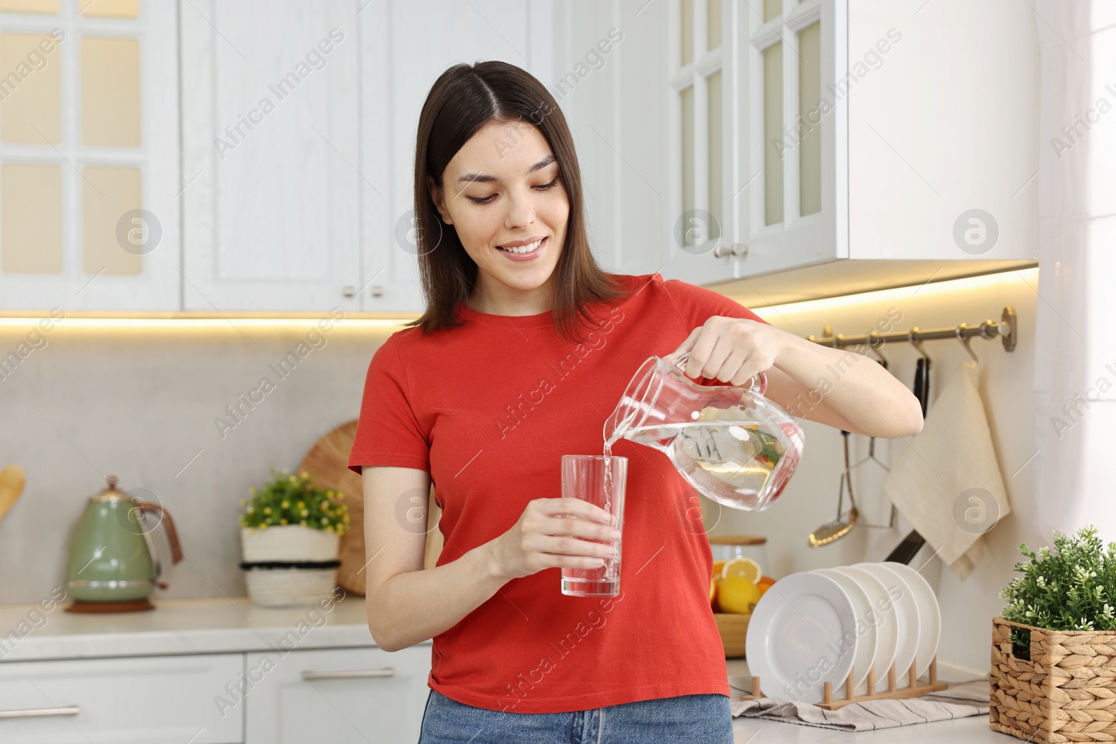 Photo of Young woman pouring water into glass from jug in kitchen