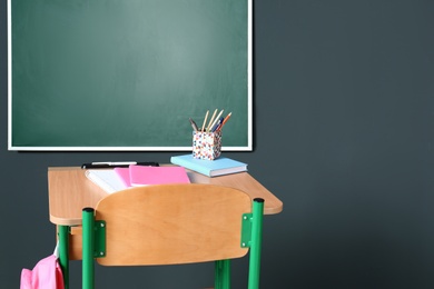 Photo of Wooden school desk with stationery and backpack near blackboard on grey wall