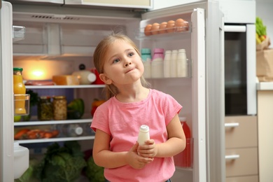 Photo of Cute little girl standing with yogurt near open refrigerator at home