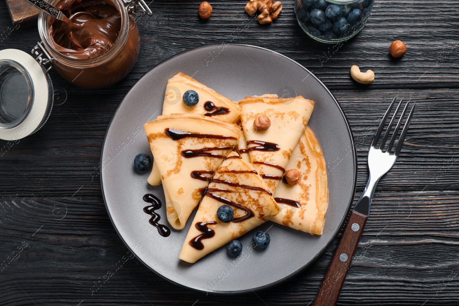 Photo of Delicious thin pancakes with chocolate, blueberries and nuts on black wooden table, flat lay