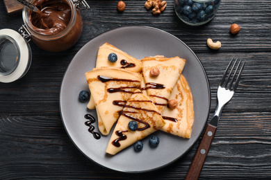 Delicious thin pancakes with chocolate, blueberries and nuts on black wooden table, flat lay