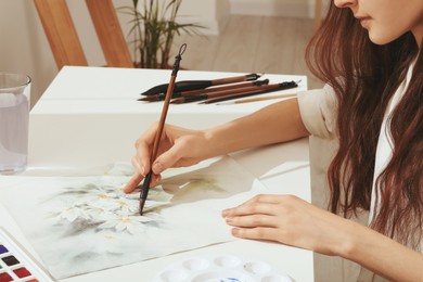 Woman painting flowers with watercolor at white table indoors, closeup. Creative artwork