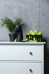 Photo of Stylish decor, basket with apples and houseplant on chest of drawers near grey wall indoors. Interior design