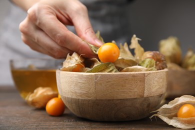 Photo of Woman peeling physalis fruit from calyxes at wooden table, closeup