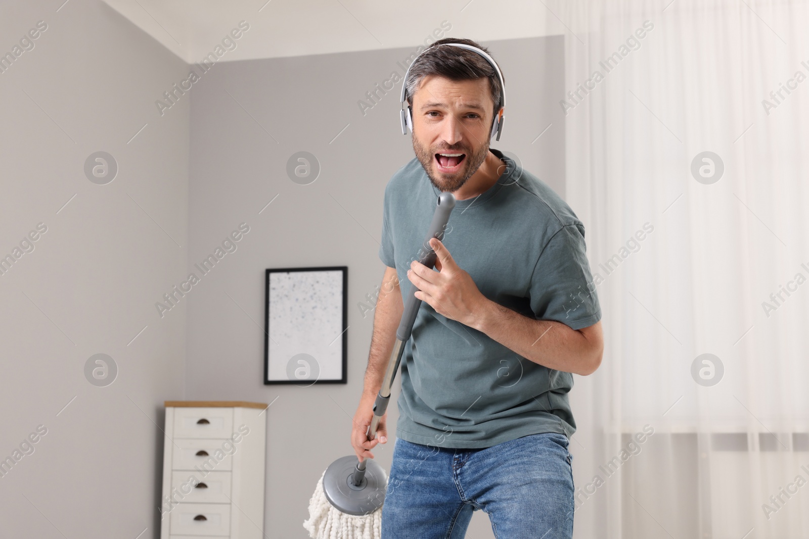 Photo of Enjoying cleaning. Happy man in headphones with mop singing while tidying up at home