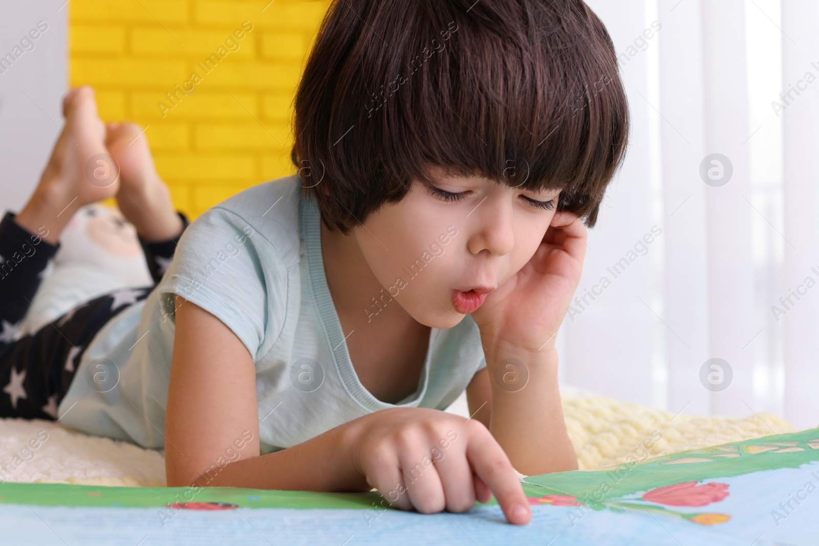 Photo of Cute little boy reading book on bed at home, closeup