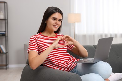 Photo of Happy young woman having video chat via laptop and making heart with hands on sofa in living room