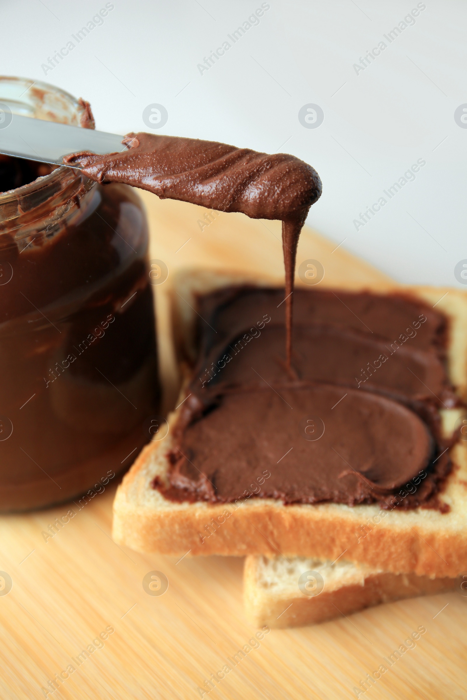 Photo of Tasty toast with chocolate paste, knife and jar on wooden board, closeup