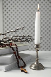 Books, burning candle, rosary beads and willow branches on white marble table, closeup