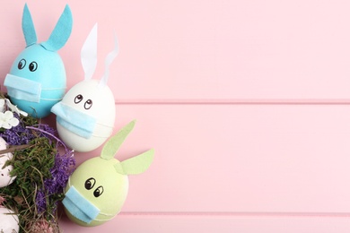 Photo of Painted eggs decorated with bunny ears, protective masks and space for text on pink wooden table, flat lay. Easter holiday during COVID-19 quarantine