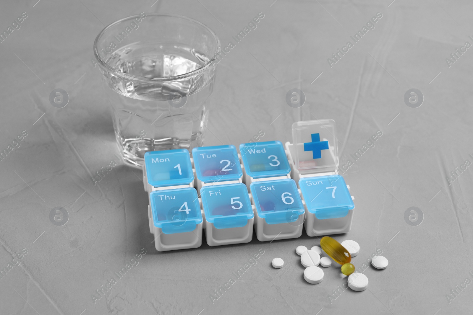 Photo of Weekly pill box with medicaments and glass of water on grey table