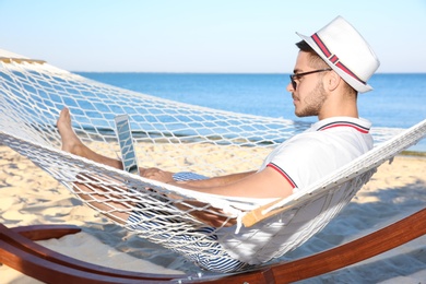 Photo of Young man with laptop resting in hammock at seaside