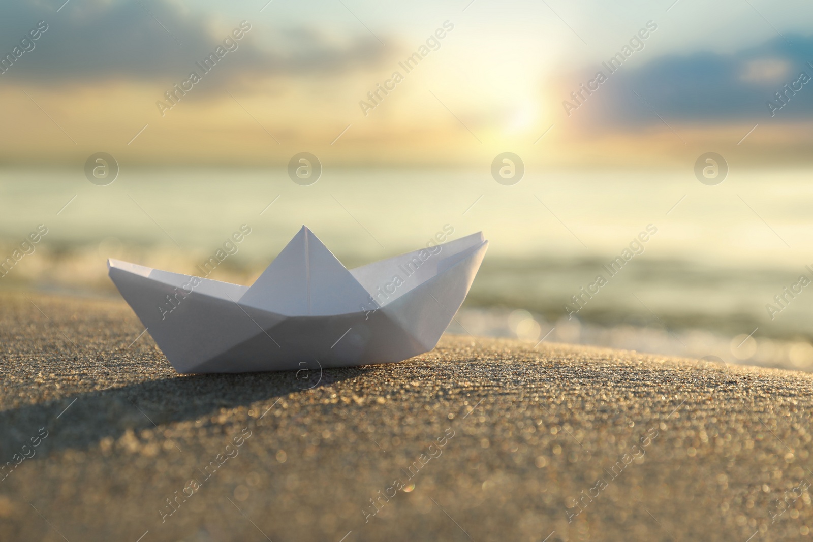 Photo of White paper boat on sand near sea at sunset, closeup. Space for text