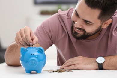 Photo of Happy young man putting money into piggy bank at white table indoors