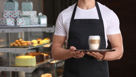 Photo of Young waiter holding tray with glass of coffee at workplace