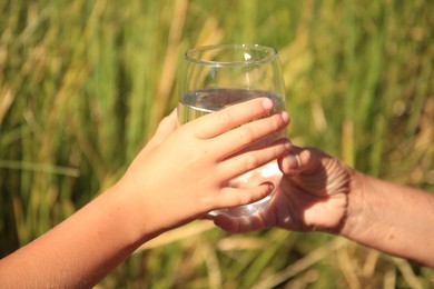 Photo of Child giving glass of water to elderly woman outdoors on sunny day, closeup