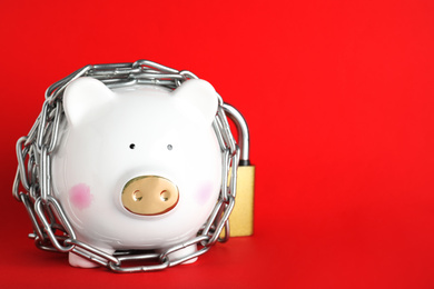 Photo of Piggy bank  with steel chain and padlock on red background, space for text. Money safety concept