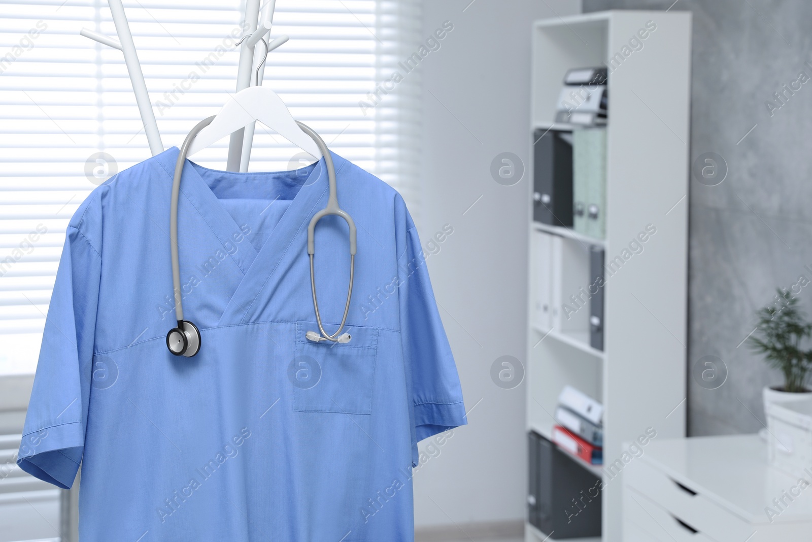 Photo of Blue medical uniform and stethoscope hanging on rack in clinic. Space for text