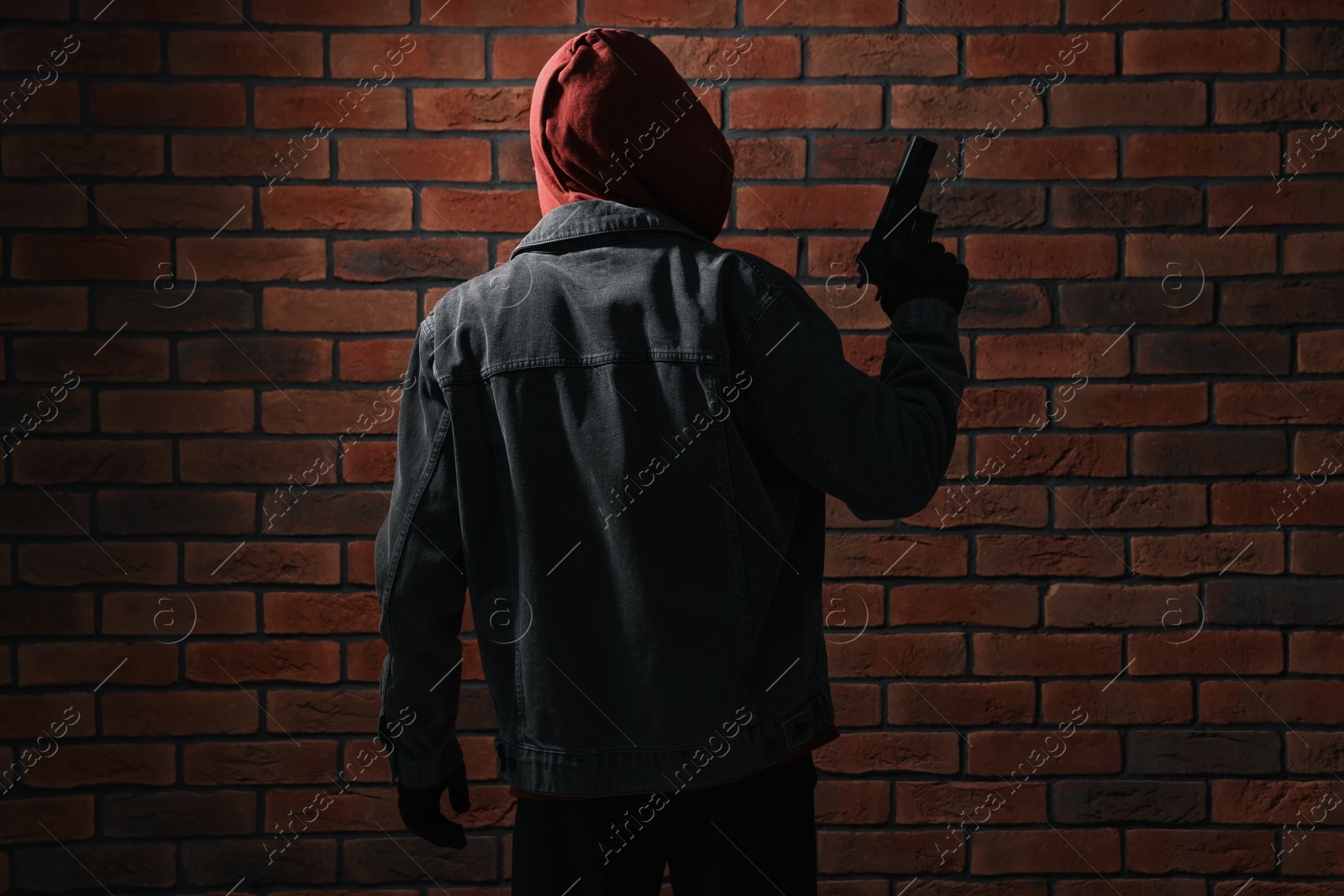 Photo of Thief in hoodie with gun against red brick wall, back view